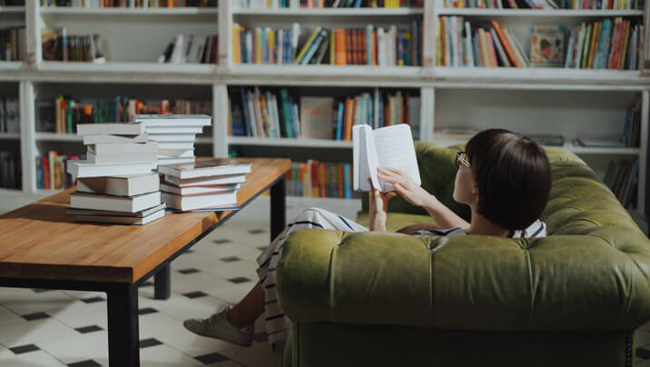 Person reading on a green sofa in a library with bookshelves.