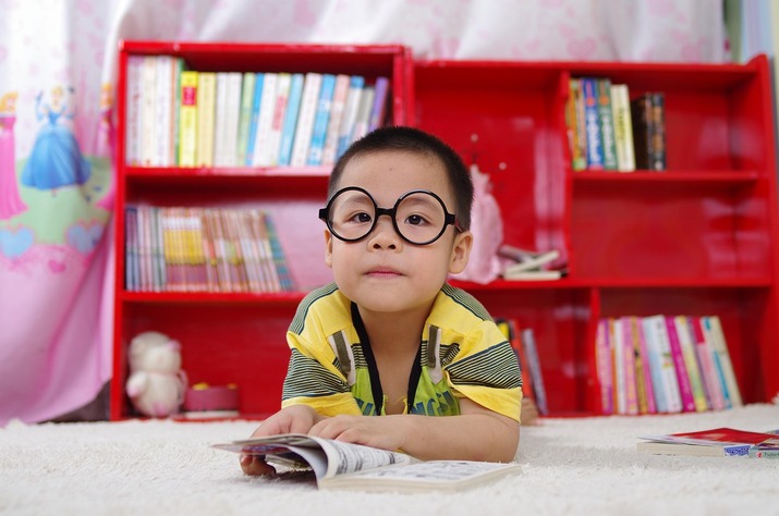 Child wearing glasses reading a book in front of a bookshelf.