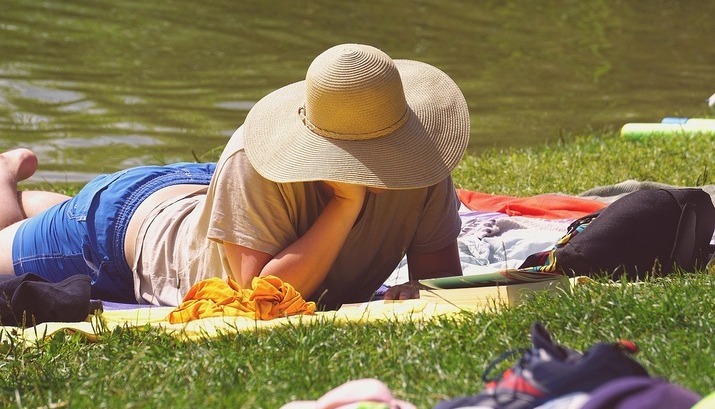 Person in large sun hat reading on a blanket by a pond.